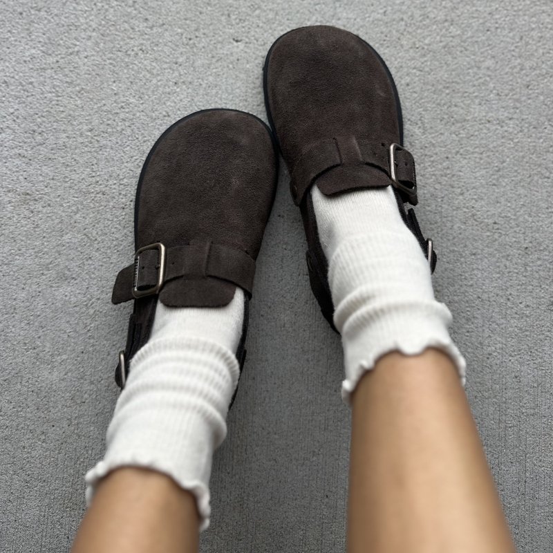 Top down look at a person's feet with their toes angled together wearing a pair of dark brown suede Barebound Ikarus II barefoot clogs. They've been paired with tall ruffled slouchy socks