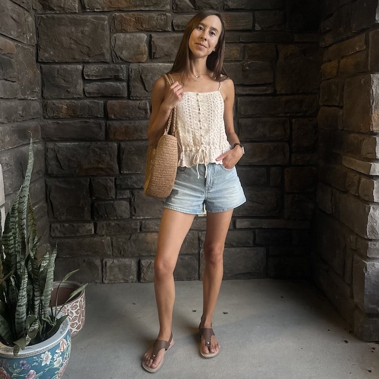 Full length photo of a woman with long straight brown hair standing in a stone alcove wearing light colored denim shorts, a crochet style cream tank, and Be Lenka Promenade 2.0 barefoot sandals