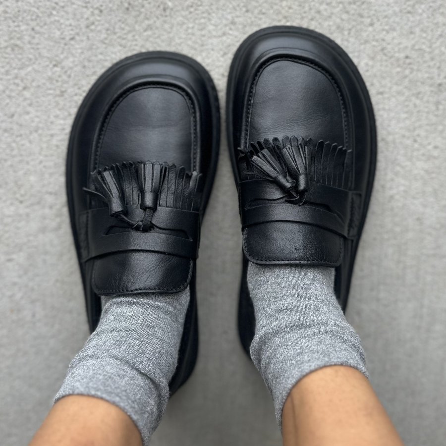 A top down close up of a pair of Fleeters Plum Loafers with fringes and tassels in full black leather. They have a wide toe box, and are worn with slouchy gray socks