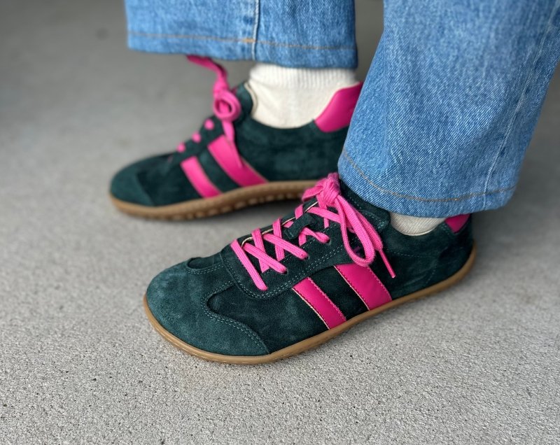 Side view of a person wearing dark green suede sneakers with bright pink stripes and laces. Their light denim pants are resting at the top of the shoe. Koel Ila Suede sneakers in green and pink shown with a wide toe box and flexible outsole