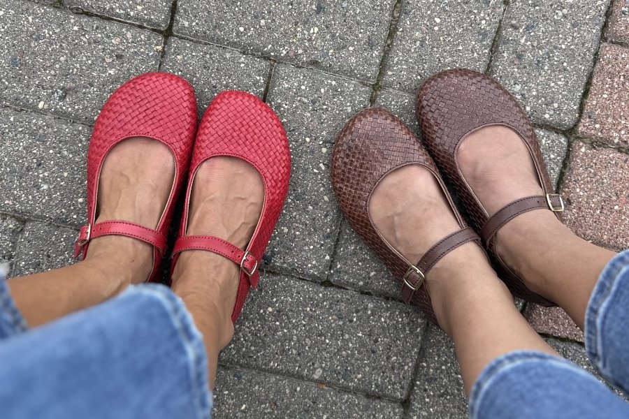 A photo of two people's feet in Paidal Woven leather mary janes with a wide toe box, shown in red leather and a dark brown chocolate leather. They're both standing on a stone walkway.