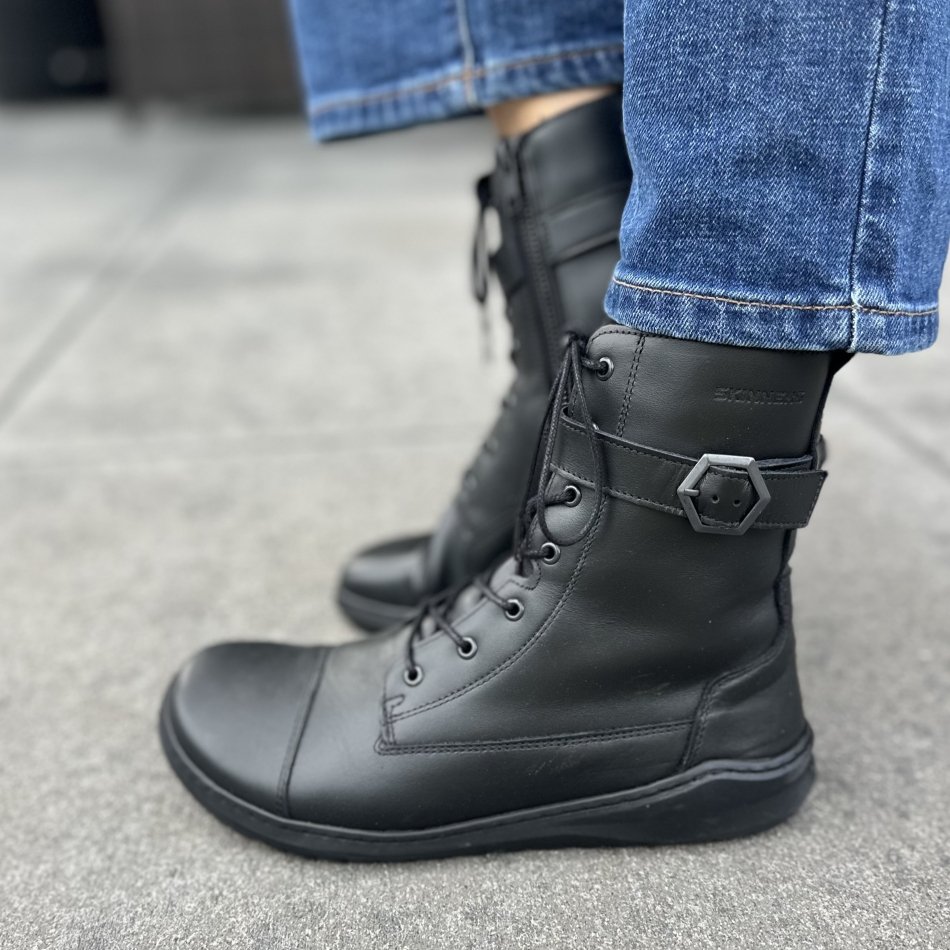 Side view of a person's feet in a pair of black leather lace up barefoot boots from Skinners with silver hexagon buckle around the top. The back foot is flexed up, and the closer foot is displaying the flat outsole. A pair of jeans are resting at the top of the boots.