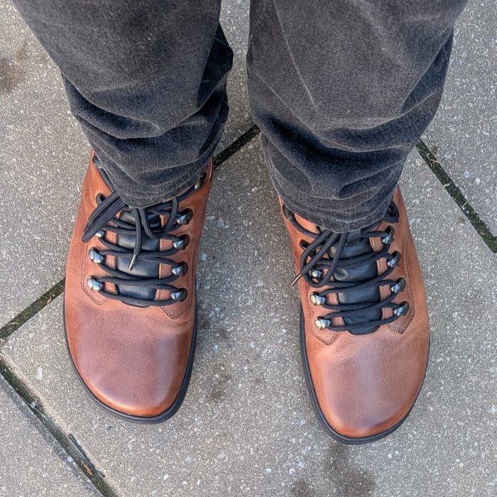Top down view of a pair of shiny new Skinners Cavalier brown leather barefoot warm boots with black laces for multi functional wear. Shown with black jeans, standing on concrete.