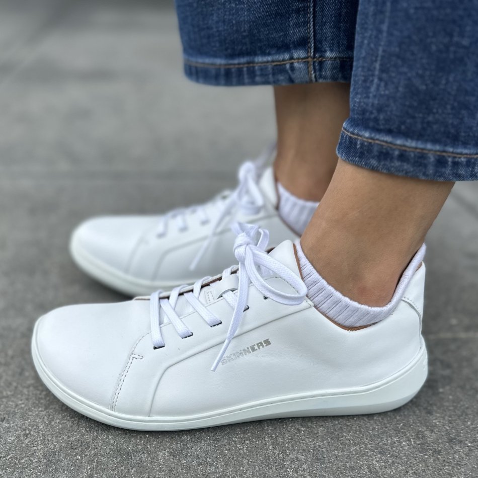 Side view of a pair of white leather barefoot Skinners sneakers with a wide toe box and flat flexible outsole. They're shown with cropped denim and white ankle socks.
