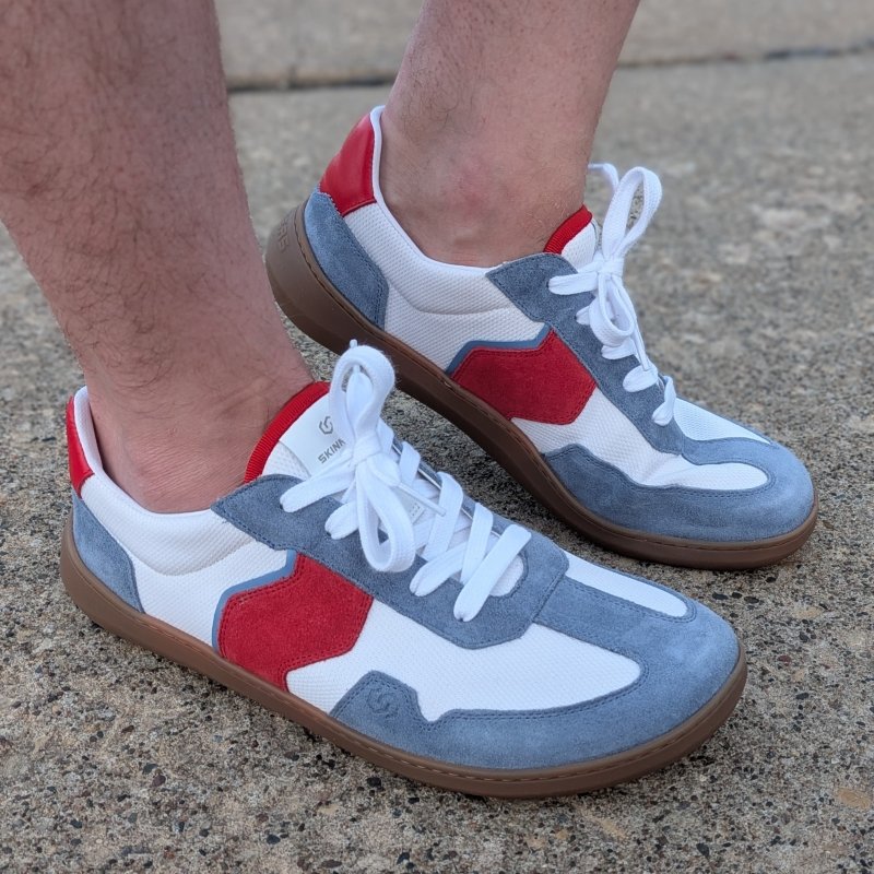 Side view of a man's feet close up wearing a pair of Skinners Timehacker barefoot sneakers in blue suede, with white and dark red accents. The back foot is lifted slightly to demonstrate flexibility of the flat sole