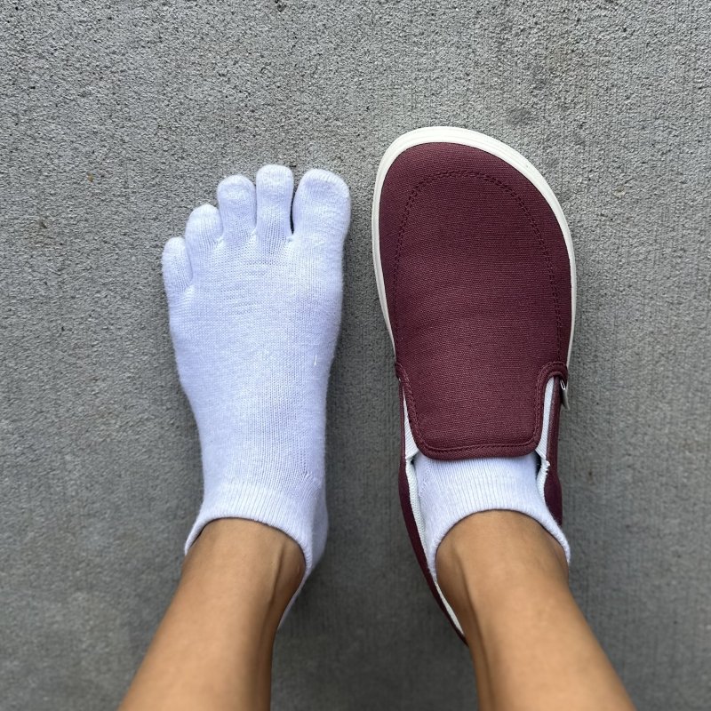 A top down close up of a person wearing one Splay Rev Slip-on sneaker in maroon canvas on one foot and a pair of white toe socks on the other foot to show how the shoe is foot shaped.