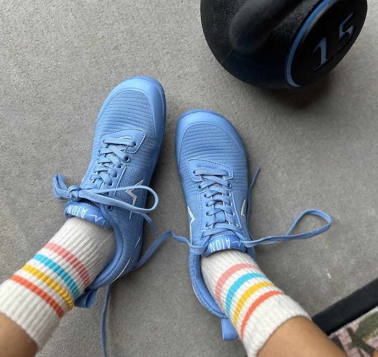 A top down view of a pair of feet resting on concrete wearing Aion barefoot training shoes in ice blue with a kettlebell resting nearby