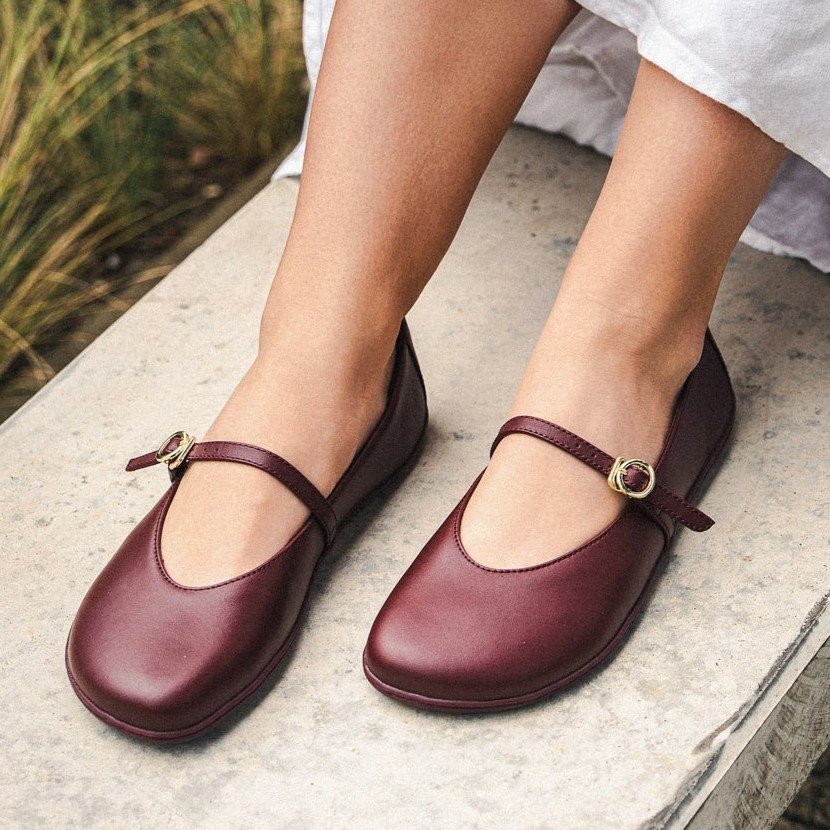 A close up view of a pair of Be Lenka Amalea Burgundy barefoot mary janes being worn with a white dress, resting on a stone wall.