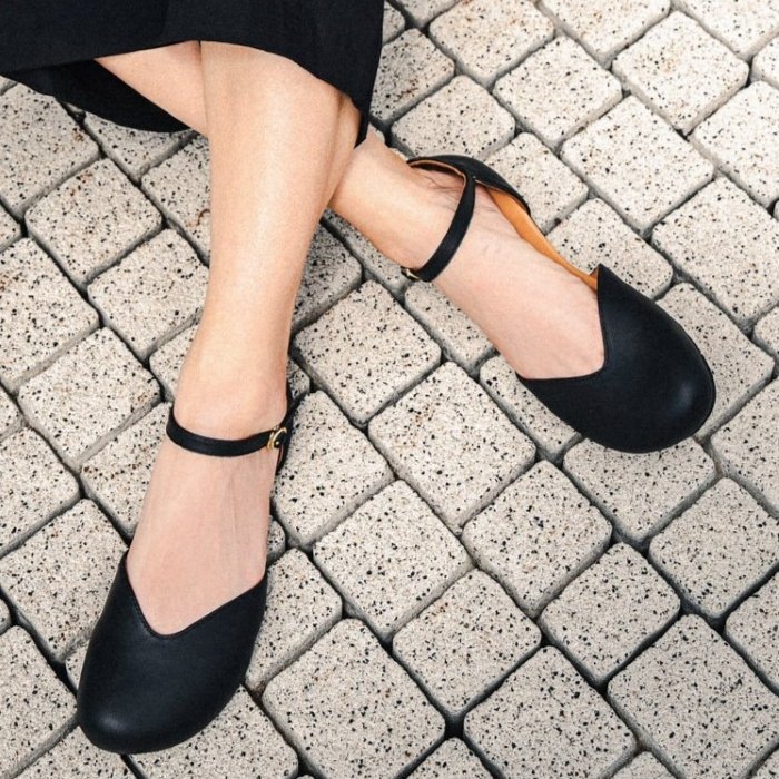 A close up of a person's feet in Be Lenka Lilea barefoot flats with a V shaped vamp. The legs are crossed and resting on a stone pathway.