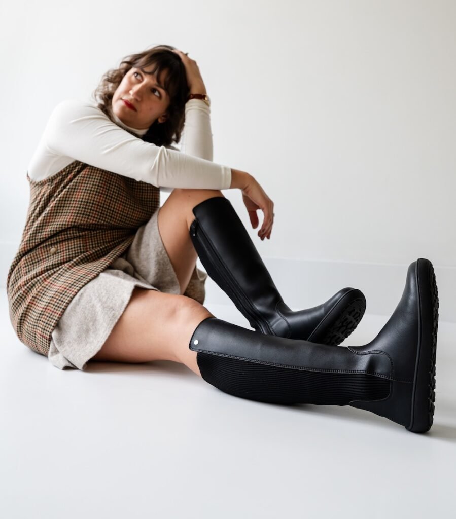 A woman sitting on the ground with her legs out in front of her, wearing a plaid wool dress and black leather Be Lenka Symphony knee high barefoot riding boots