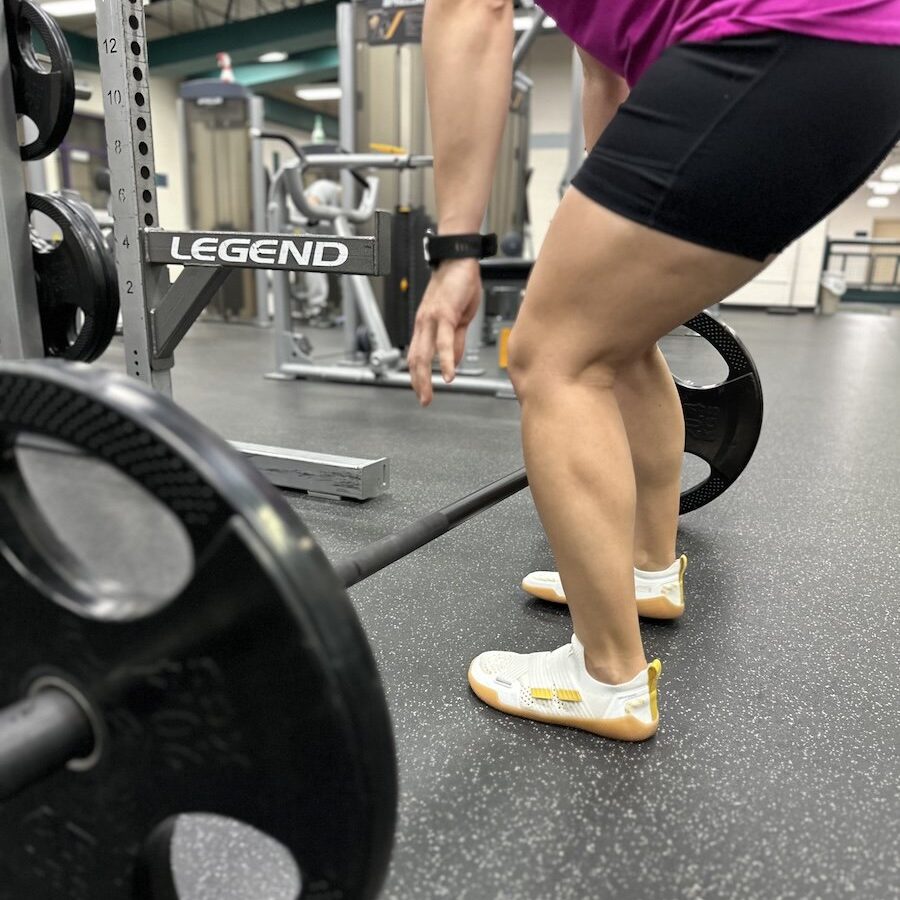 A woman bending over to pick up a barbell for a deadlift, weightlifting in the gym wearing TYR DropZero barefoot trainers
