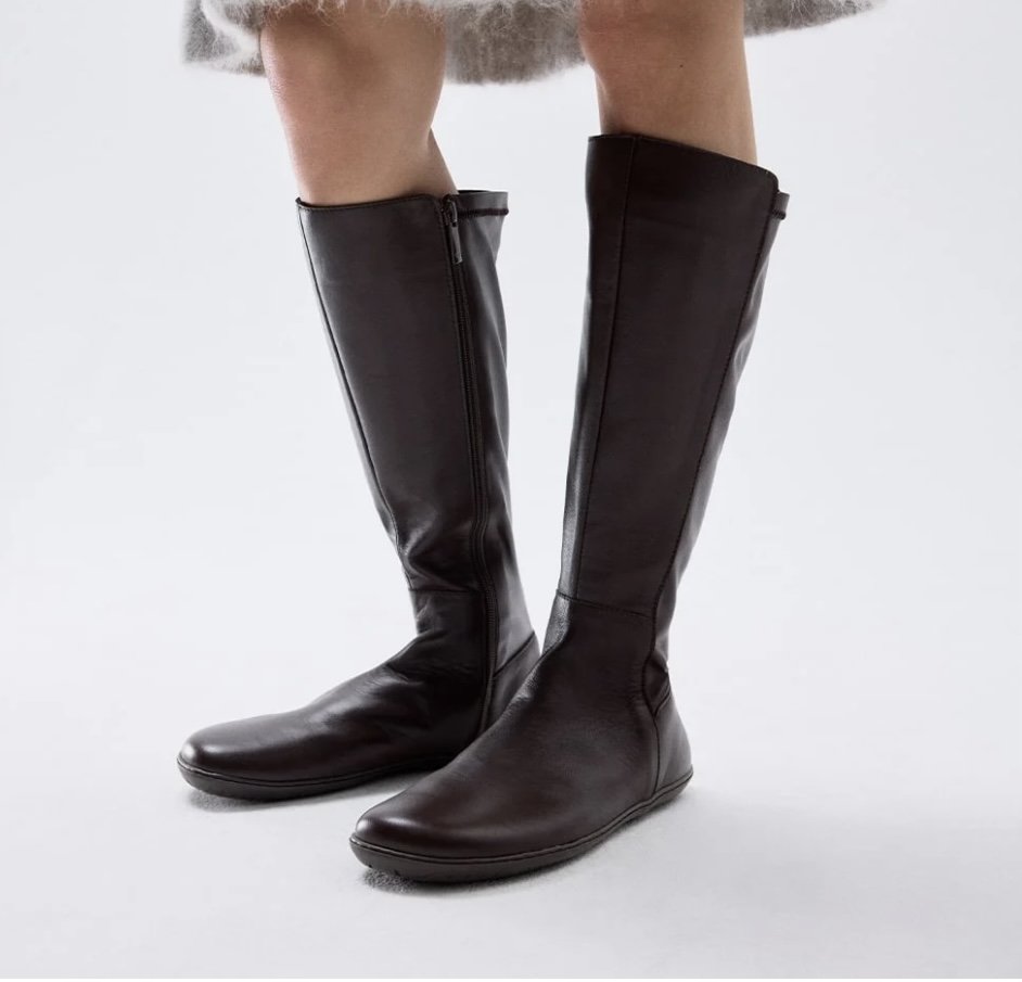 A close up side view of a woman's legs from the knee down wearing Groundies barefoot tall leather boots