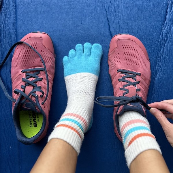 A top down view of a pair of feet resting on an exercise mat. One foot is bare with a toe sock on, the other foot is wearing a wide toe box barefoot minimalist Inov8 Bare gym shoes