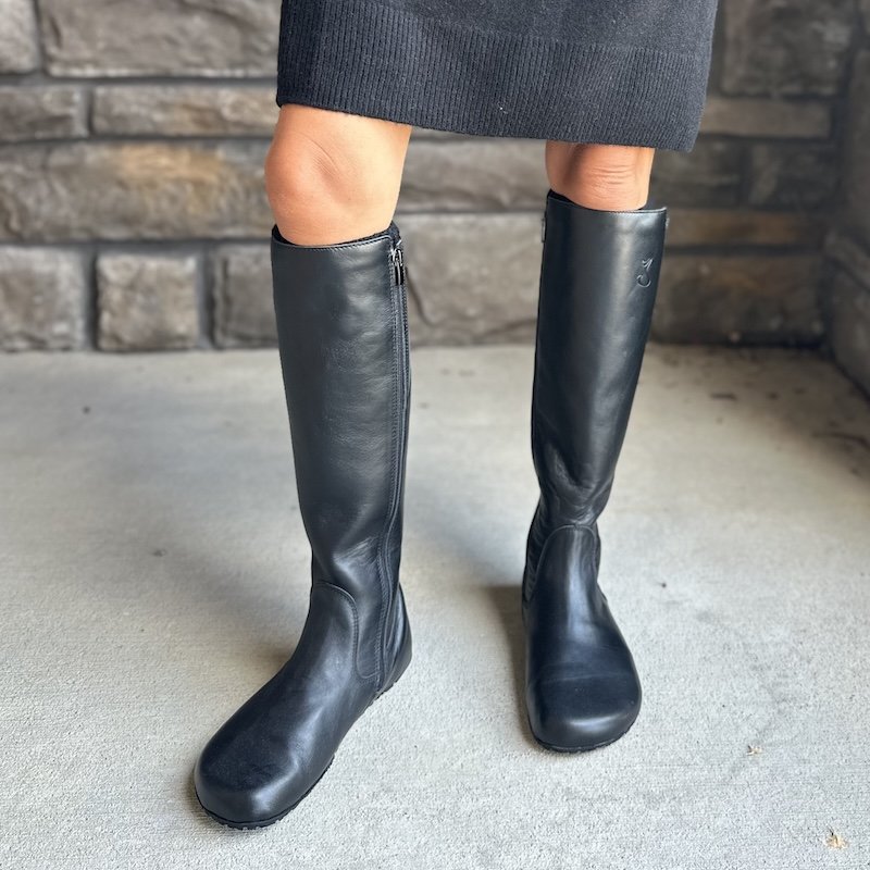 A close up view of a woman's legs from the thighs down standing on concrete wearing black leather fleece lined Jenon Leather Eternal high wide toe box barefoot riding boots