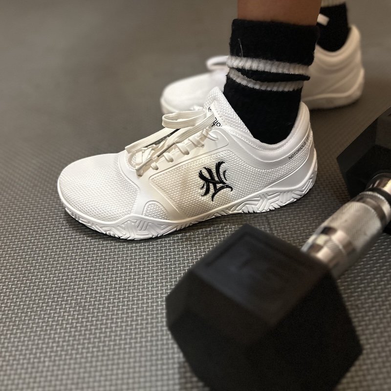 A pair of feet on an exercise mat wearing black socks and white Notorious Lift Radix Pro wide toe box lifting shoes, with a dumbbell next them