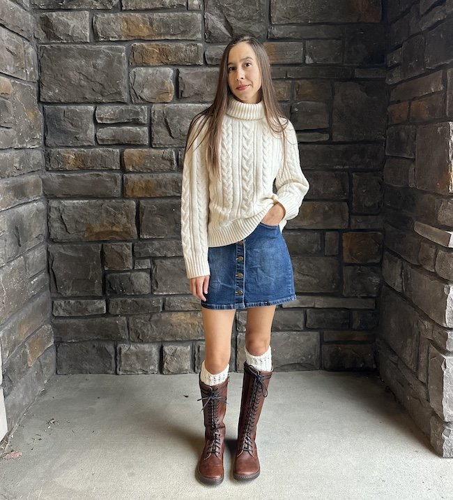 A woman standing in front of a stone wall smiling at the camera wearing a chunky sweater, denim skirt, and knee high lace up barefoot wide toe box boots in brown leather - the Peerko Empire Brandy