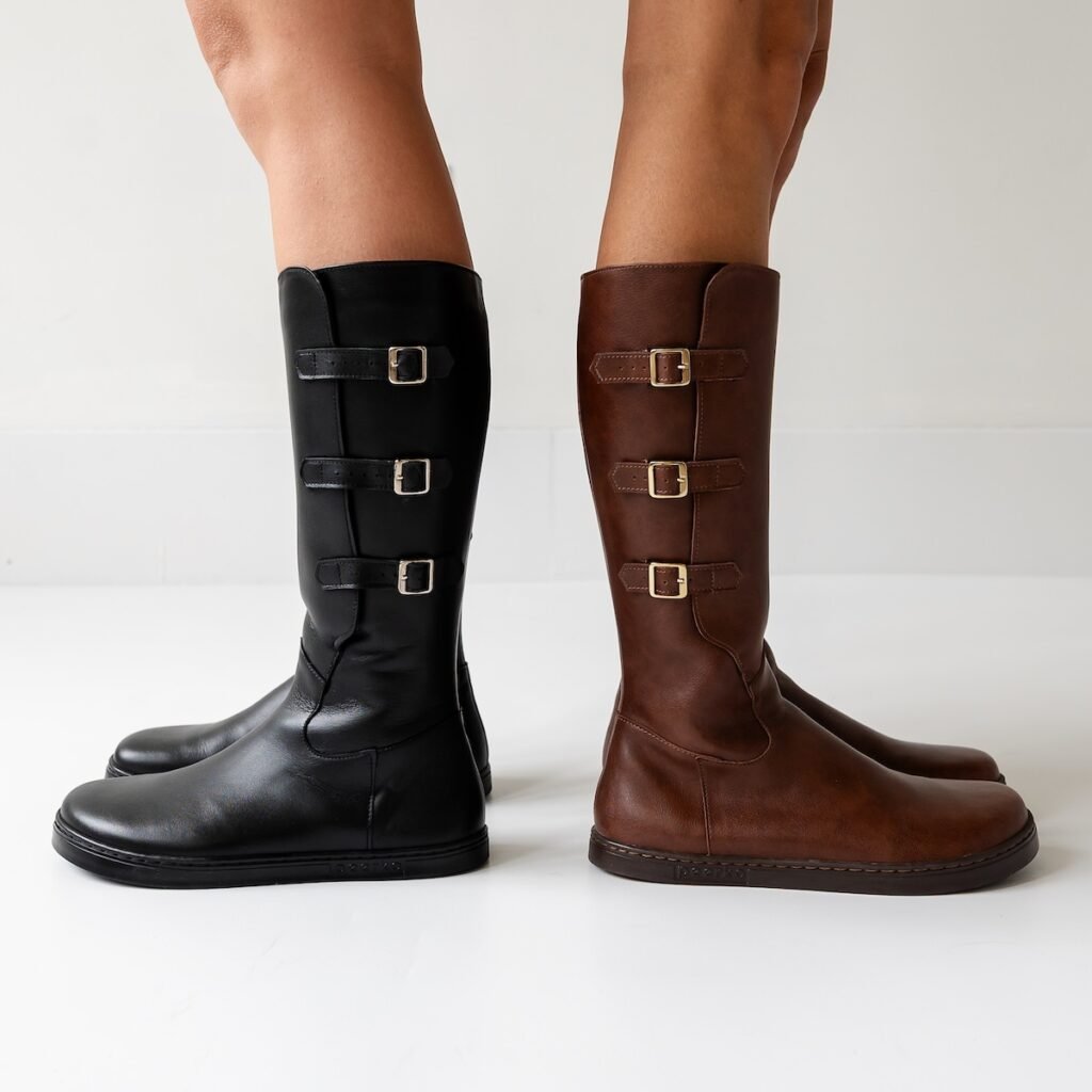 A close up of two women's legs, standing back to back from the knees down wearing Peerko Joana knee high barefoot wide toe box boots with adjustable buckles. The left woman is wearing black leather, and the right woman is wearing brown leather.