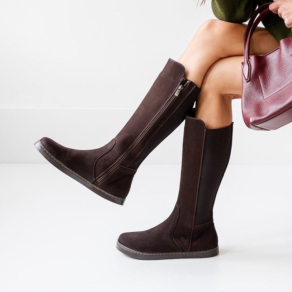 A close up of a woman's legs, sitting crossed, from the thighs down. On her feet are Peerko Regina II brown suede barefoot riding boots