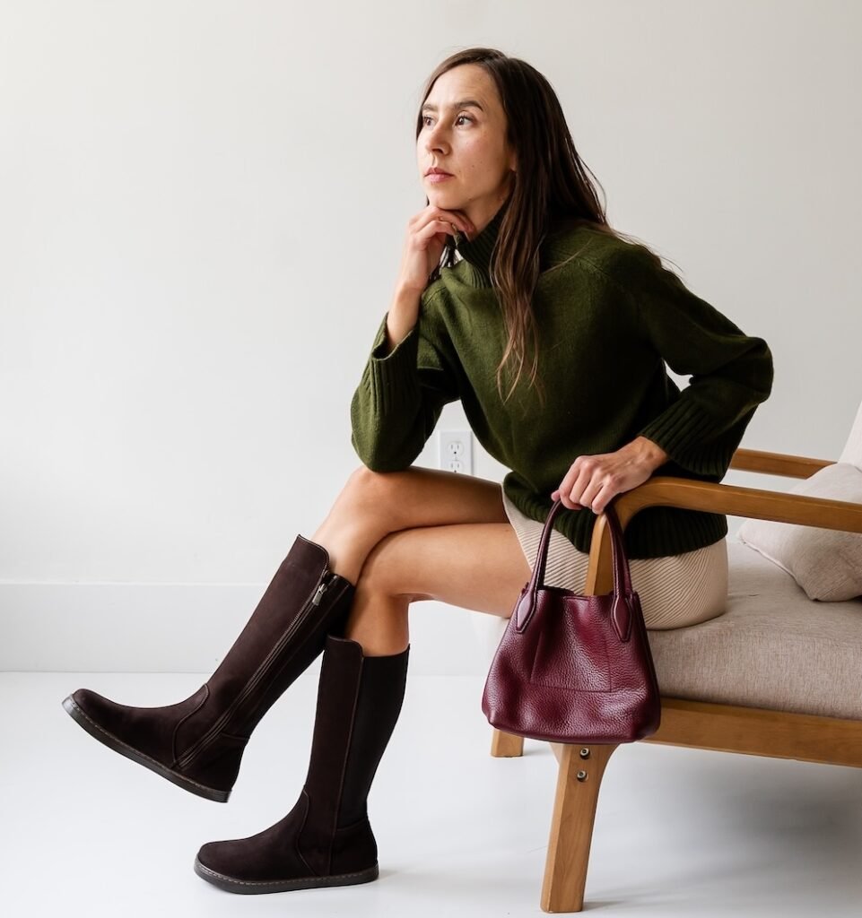 A woman sitting on a chair looking off in the distance with her legs crossed, wearing a tan skirt and green sweater. On her legs are a pair of brown suede Peerko Regina II barefoot knee high riding boots