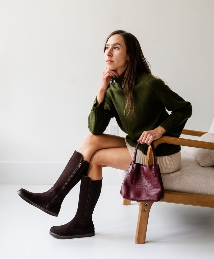 A woman sitting on a chair looking off in the distance with her legs crossed, wearing a tan skirt and green sweater. On her legs are a pair of brown suede Peerko Regina II barefoot knee high riding boots