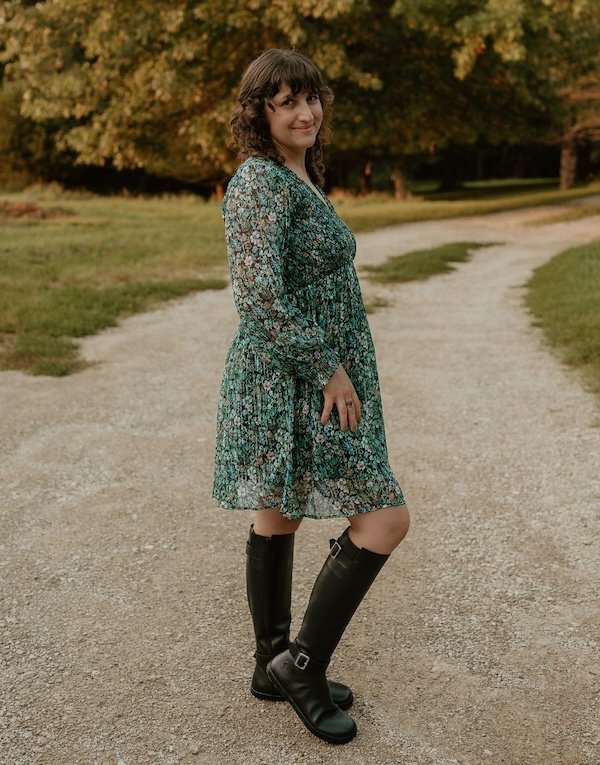 A woman standing in a dirt road smiling at the camera, wearing a floral mini dress and black knee high Shapen Glam barefoot riding boots with metal buckles