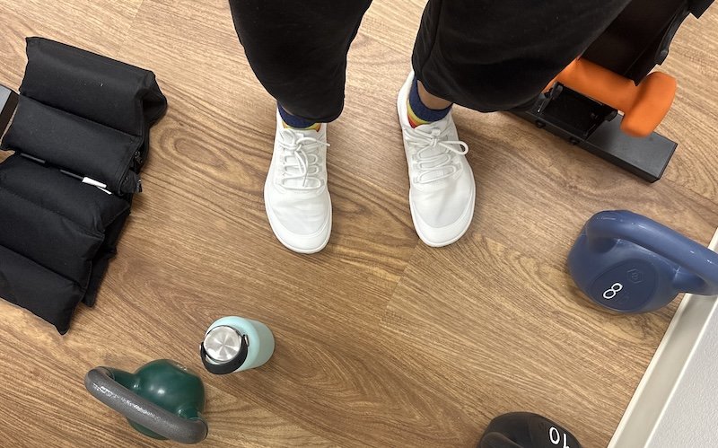 Top down image of a person's feet surrounded by exercise equipment. On her feet are Tolos Archetype barefoot gym sneakers with a wide toe box in white.