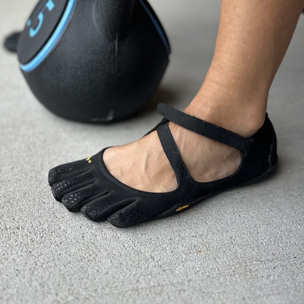 A close up side view of a single foot planted on the ground next to a kettlebell wearing Vibram Five Fingers V soul barefoot shoes