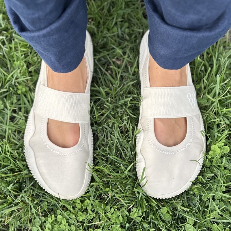 Top down view of a woman's feet in Vivobarefoot Motus Studio Ballet sporty flats with an elastic strap. They are standing on grassy ground with blue linen pants hanging near the ankle.