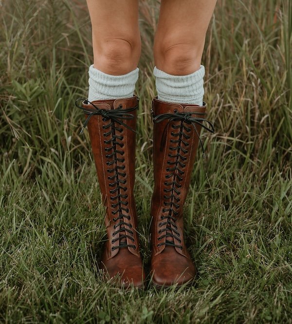 A close up of a woman's leg's visible from mid though down, standing in front of tall grass wearing knee high boots and socks. The boots are a cognac brown, tall lace up boots - the Peerko Emplor leatherire in Brandy co