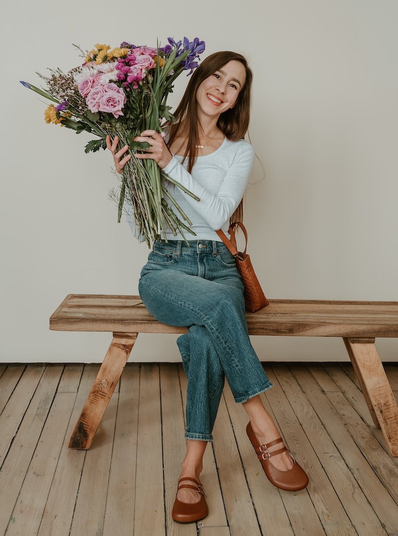 Full body view of a woman sitting on a wooden bench holding a large bouquet of flowers. She has on a pair of medium brown leather Jenon Leather Villet double strap barefoot mary janes.