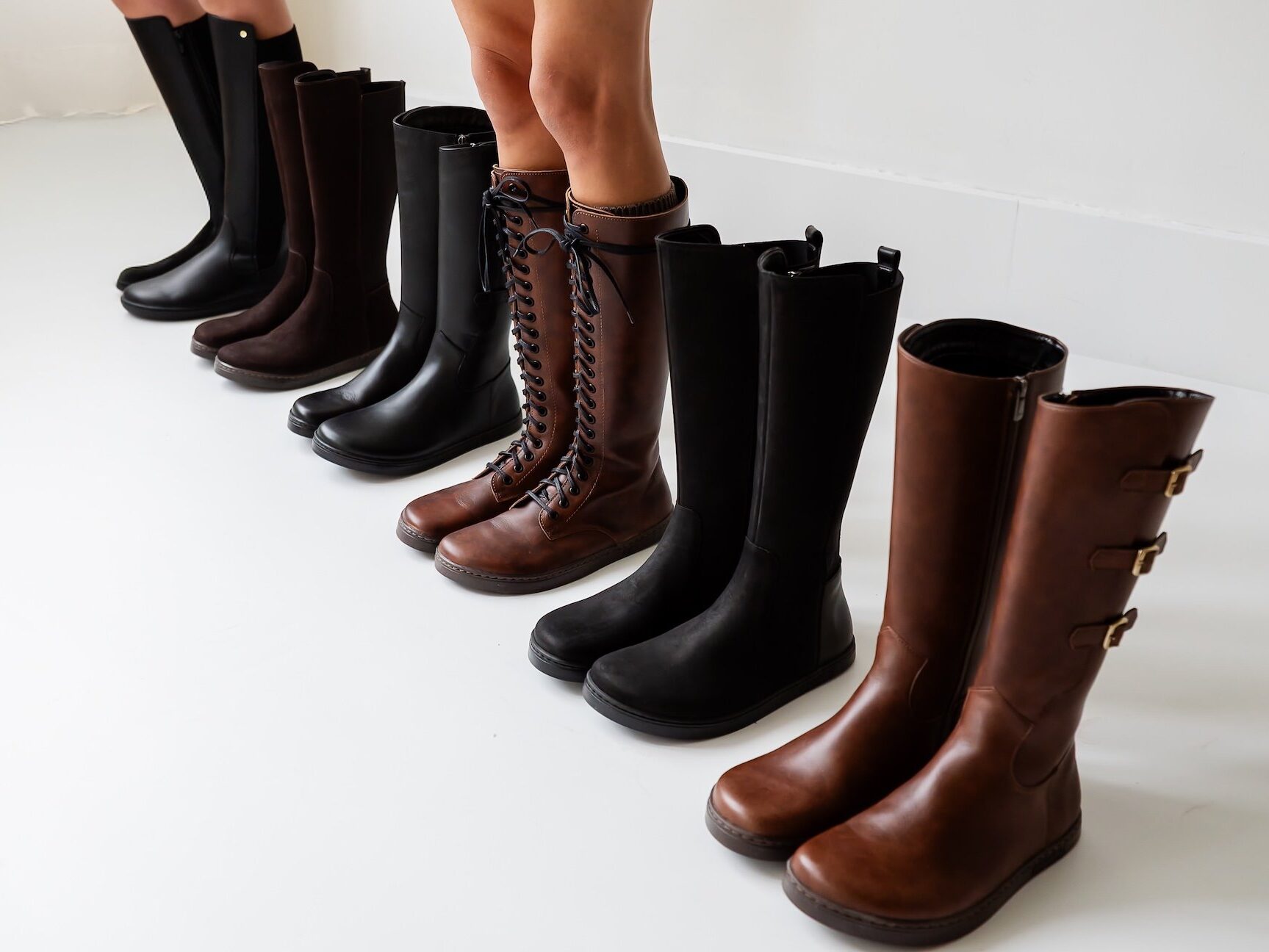 A line up 6 different barefoot knee high boots in black and brown lined up in a row
