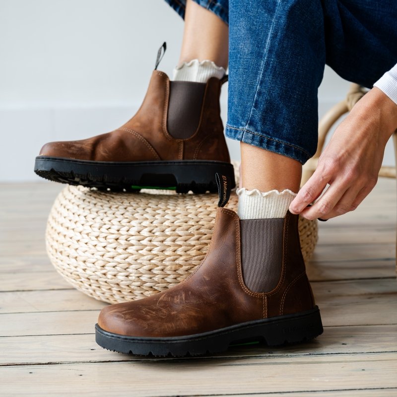 A person putting on a pair of El Naturalista barefoot Chelsea boots in brown leather. The back foot is propped up on a woven pouf. The minimalist chelsea boots are being styled with ruffled socks and dark denim.