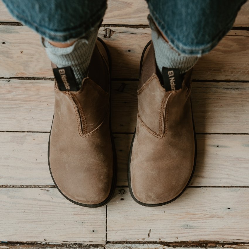 Top view of a pair of feet in barefoot El Naturalista Nomada Chelsea boots in Stone colored leather worn with ruffled socks and medium wash denim.