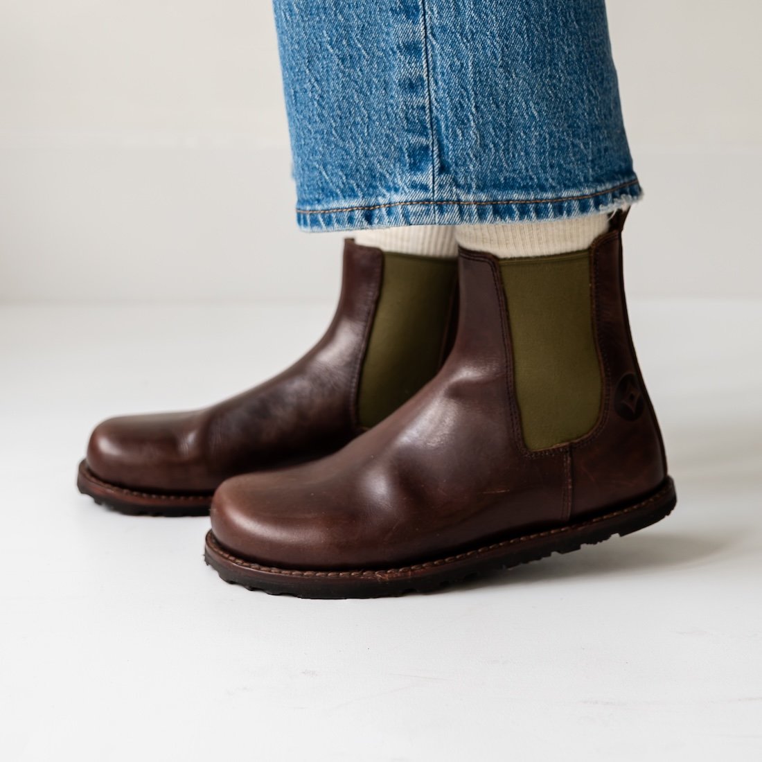 A close up side view of a person's feet in Gaucho Ninja Barefoot Chelsea boots with Goodyear Welt construction, a wide toe box, and glossy brown leather. Cream socks are visible over the top of the boot, and a pair of midtone jeans hangs just above the ankle.