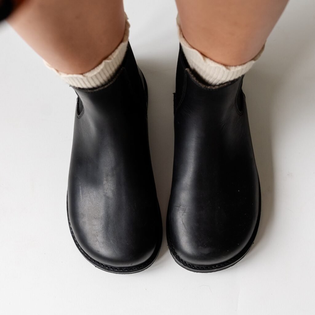 A top down view of a person's feet in Gaucho Ninja Chelsea boots in black leather with a wide toe box and flat outsole. A pair of ruffled cream Tabio socks are poking out the top of the boots.