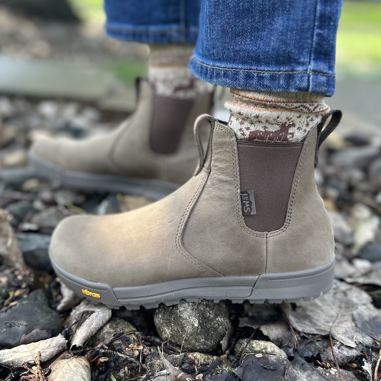 A close up side view of a pair of feet standing on rocks outside wearing wool socks and Lems Chelsea Tuff Pro wide toe box boots in sand leather, from the side