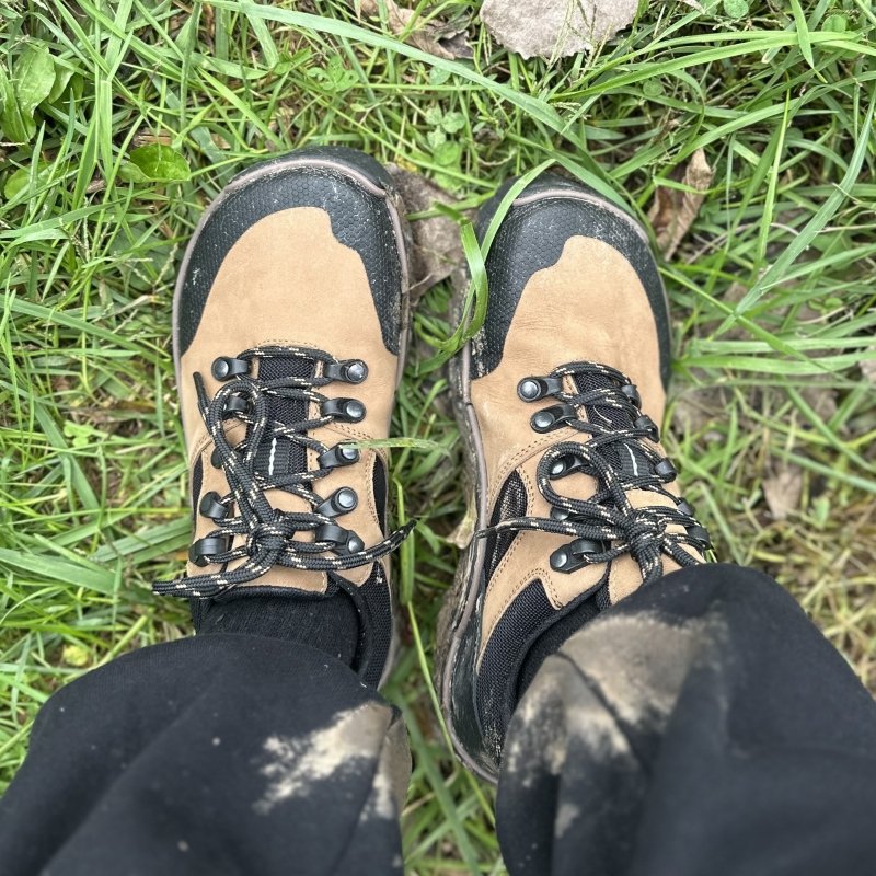 Top down view of a person in muddy hiking shoes and pants. The Magical Shoes Tundra low top hiking shoes in tan, with a reinforced wide toe box, and flexible outsole