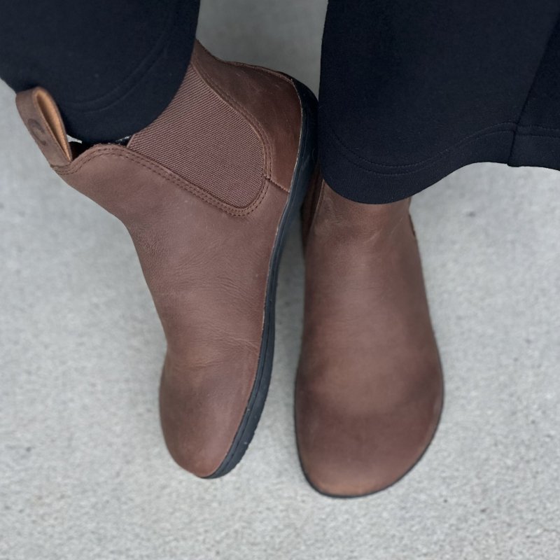 Top down and side view of a person in brown leather chelsea boots from ZuZu. Black pants are hanging near the ankle, and the wide toe box is displayed along with the zero drop outsole.