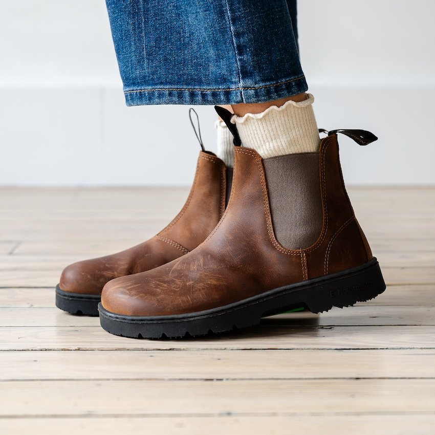 A side view of a person's feet close up in a pair of El Naturalista minimalist chelsea boots in dark brown leather. A pair of cream ruffled socks are visible over the ankle of the boot, as well as a pair of dark jeans.