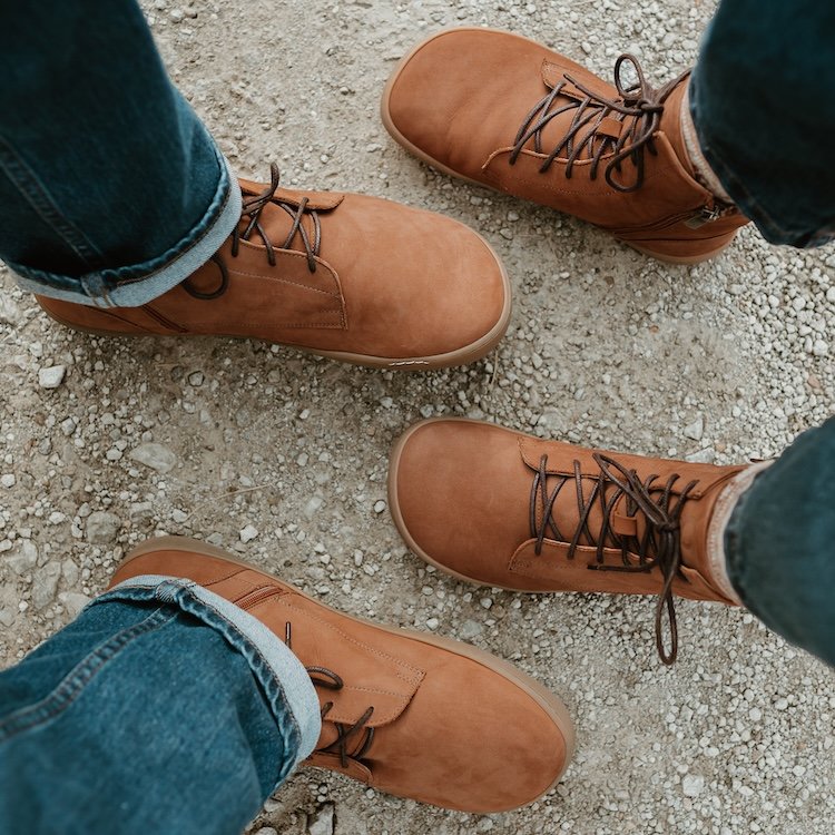 A top down view of two pairs of feet resting on dirt ground wearing denim and brown Shapen Cozy wool lined wide toe box barefoot boots.