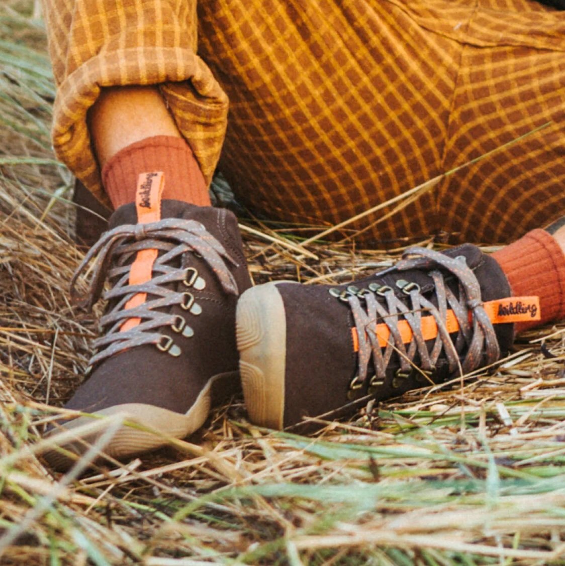A close up of a pair of feet resting on straw wearing Wildling Shoes Tejo Chocolate barefoot lace up boots with a wool lining