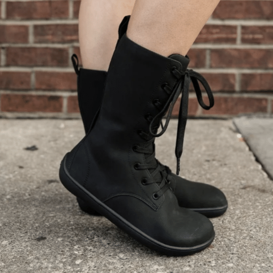 Side view of a pair of Be Lenka Evon barefoot lace up mid-height black boots. The front foot is popped up slightly showing off the zero drop outsole