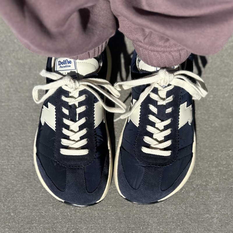 Top down view of a person's feet in mauve sweatpants and a pair of Dolfie Paradise wide toe box cushioned sneakers. The Supersonic model is shown in navy blue with a white bolt on the side and matching white laces.
