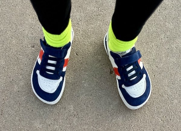 A top down view of a child's feet standing on pavement wearing neon green socks and wide toe box kids barefoot sneakers from Dolfie Paradise made of white leather with navy blue and orange accents and a velcro strap