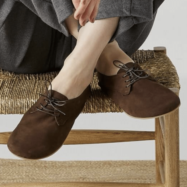 Close up of a person's feet crossed on a chair wearing a pair of Origo Derby shoes in chocolate suede.