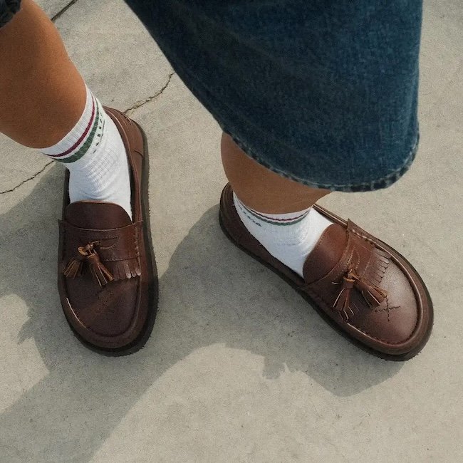 A top down view of a pair of feet standing on concrete wearing jorts and brown leather Dolfie Paradise Deck wide toe box barefoot loafers with tassels and fringe